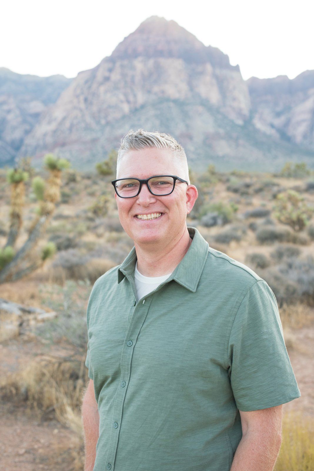 a man wearing glasses and a green shirt smiles in front of a mountain