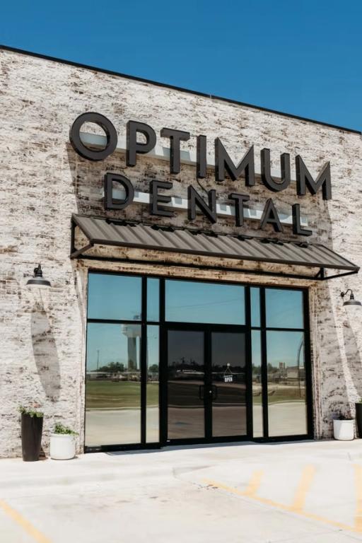 Exterior view of the Optimum Dental building with a white brick facade, black sign, and large glass entrance doors.
