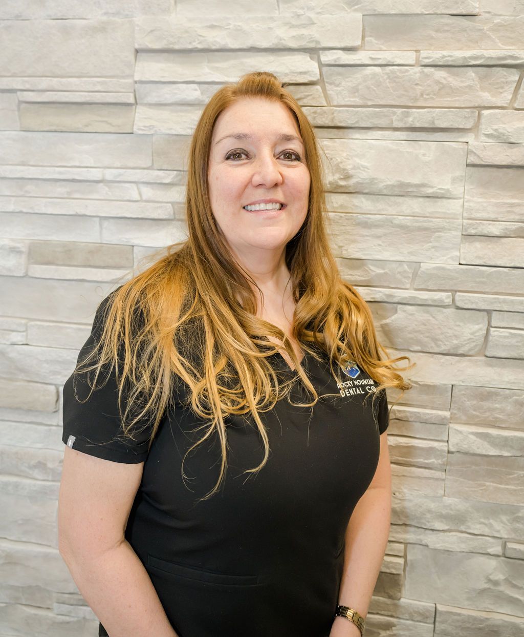 a woman wearing a black dental scrub is smiling in front of a stone wall