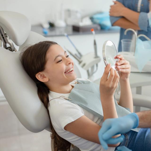 a little girl is sitting in a dental chair looking at her teeth in a mirror .