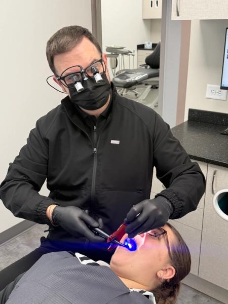 A masked dentist wearing loupes uses a blue light to treat a patient's teeth.
