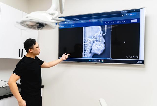 Dentist pointing at a lateral cephalometric X-ray on a large monitor.