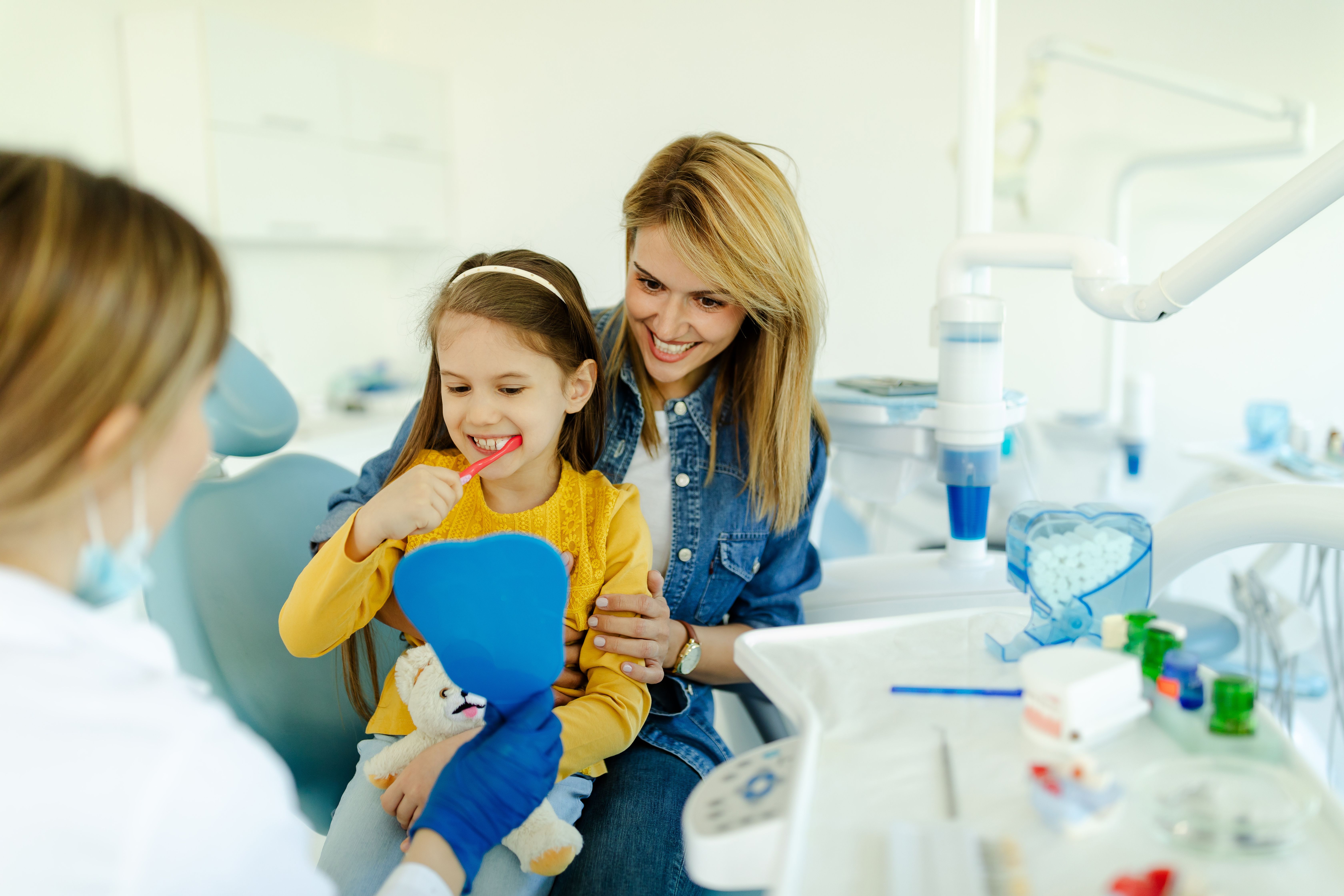 Young girl brushing teeth in a dentist's office with her mother and a dental professional.