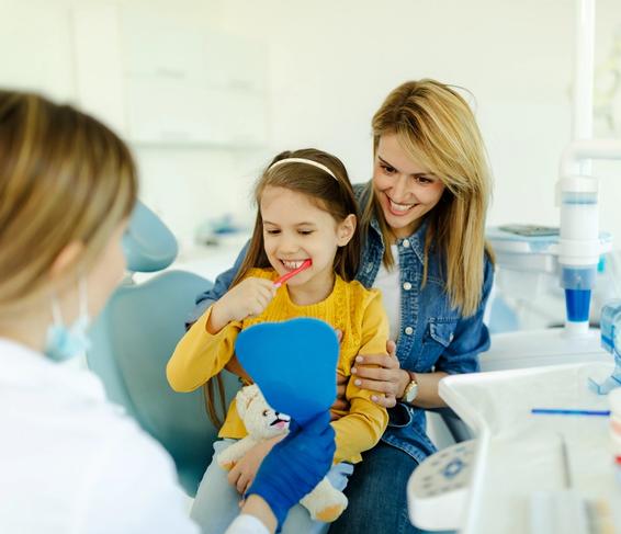 Young girl brushing teeth in a dentist's office with her mother and a dental professional.