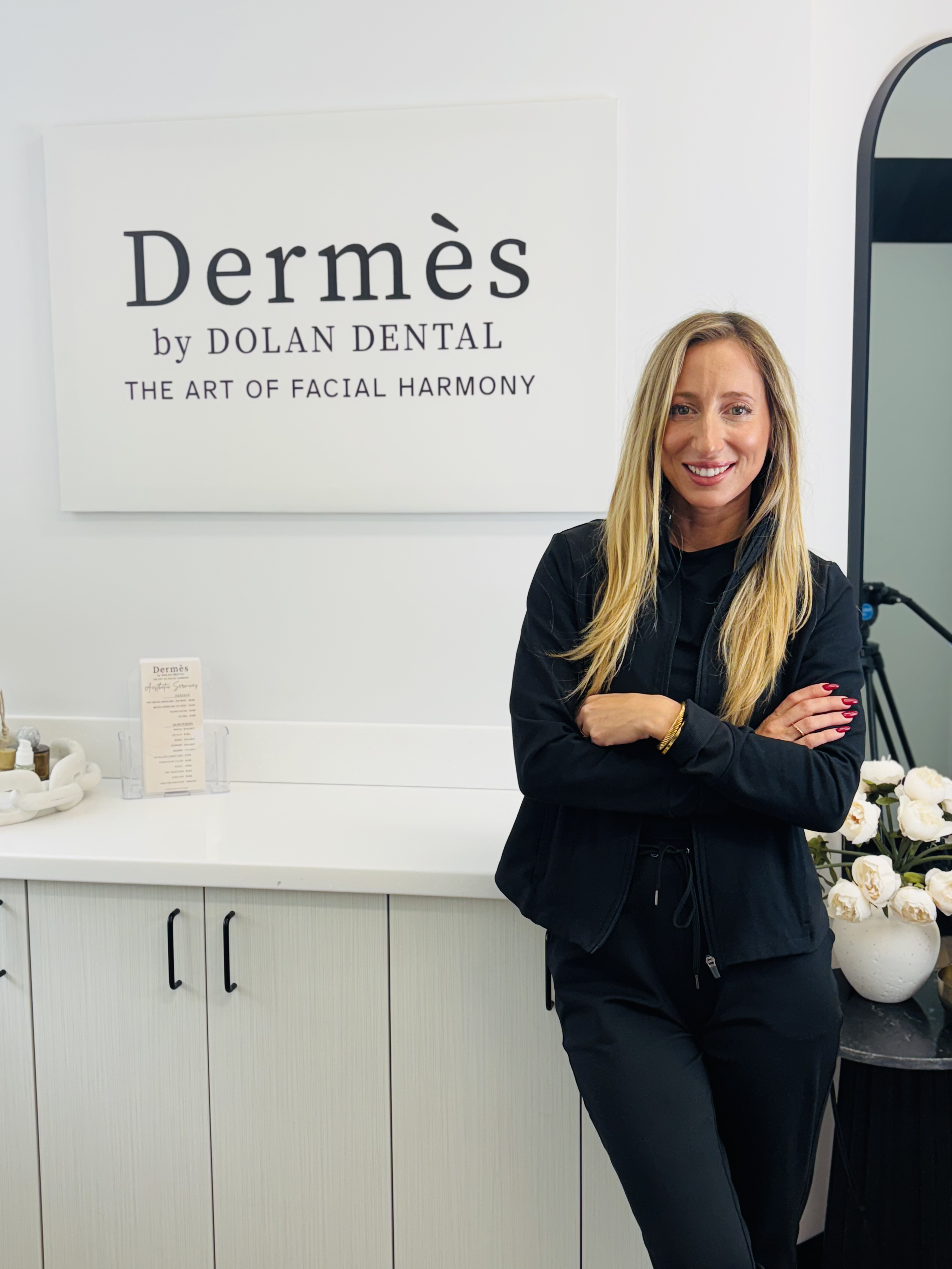 A smiling woman in black scrubs stands with crossed arms in front of a white wall with a "Dermès by Dolan Dental" sign.