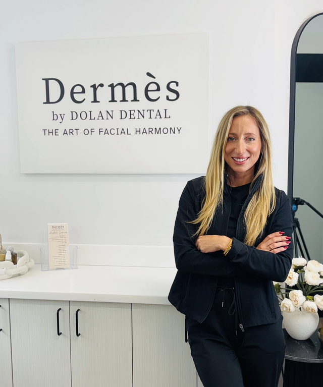 A smiling woman in black scrubs stands with crossed arms in front of a white wall with a "Dermès by Dolan Dental" sign.