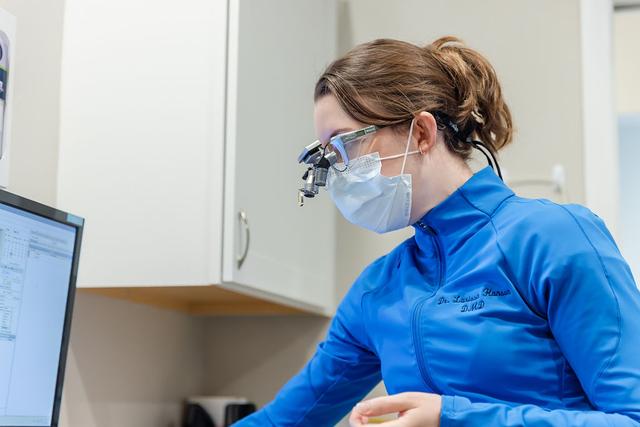 Dr. Larissa Hanson D.M.D. in a blue jacket, mask, and dental loupes, looks at a computer screen.