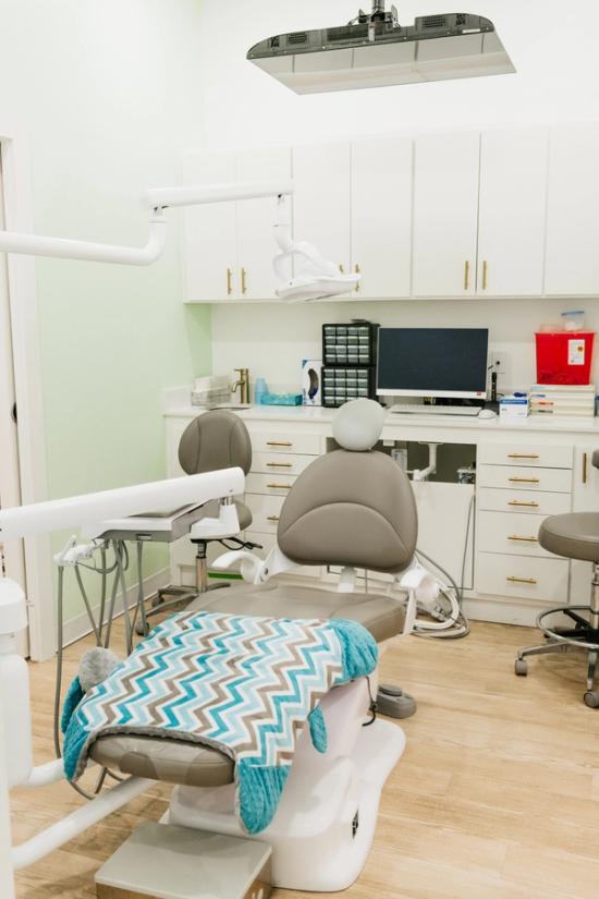 A modern dental examination room with a grey chair, chevron blanket, white cabinets, and dental equipment.
