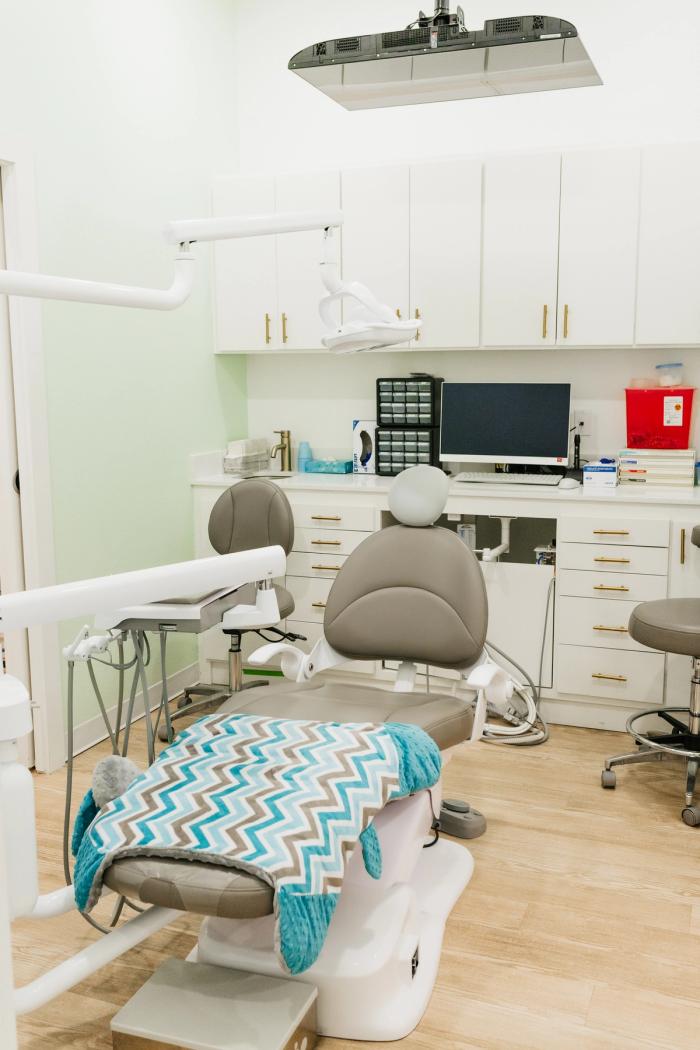 A clean dental examination room with a grey chair covered by a chevron blanket, medical equipment, and white cabinets.