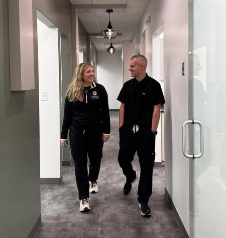 A man and a woman in black scrubs walk and smile at each other in a modern hallway.