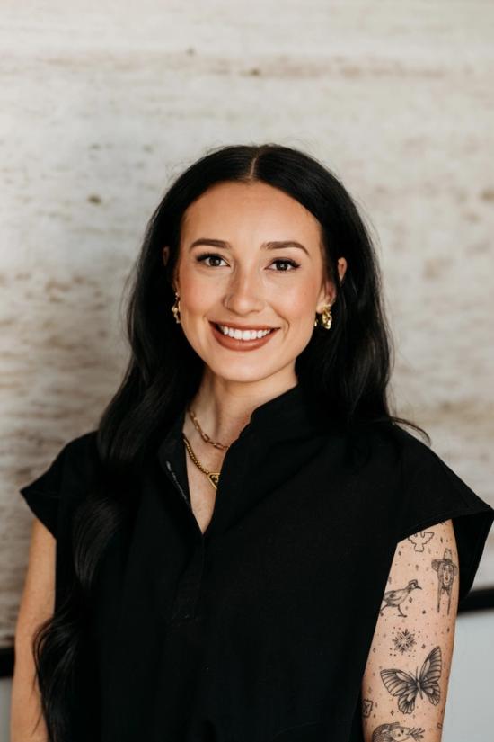 A smiling woman with long dark hair, wearing a black collared shirt, gold jewelry, and sleeve tattoos on her left arm.