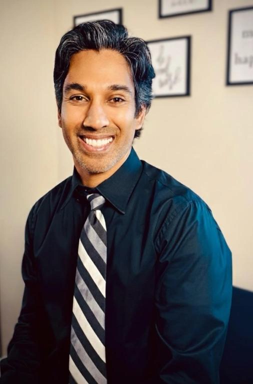 Smiling man with dark, graying hair, wearing a dark shirt and striped tie.