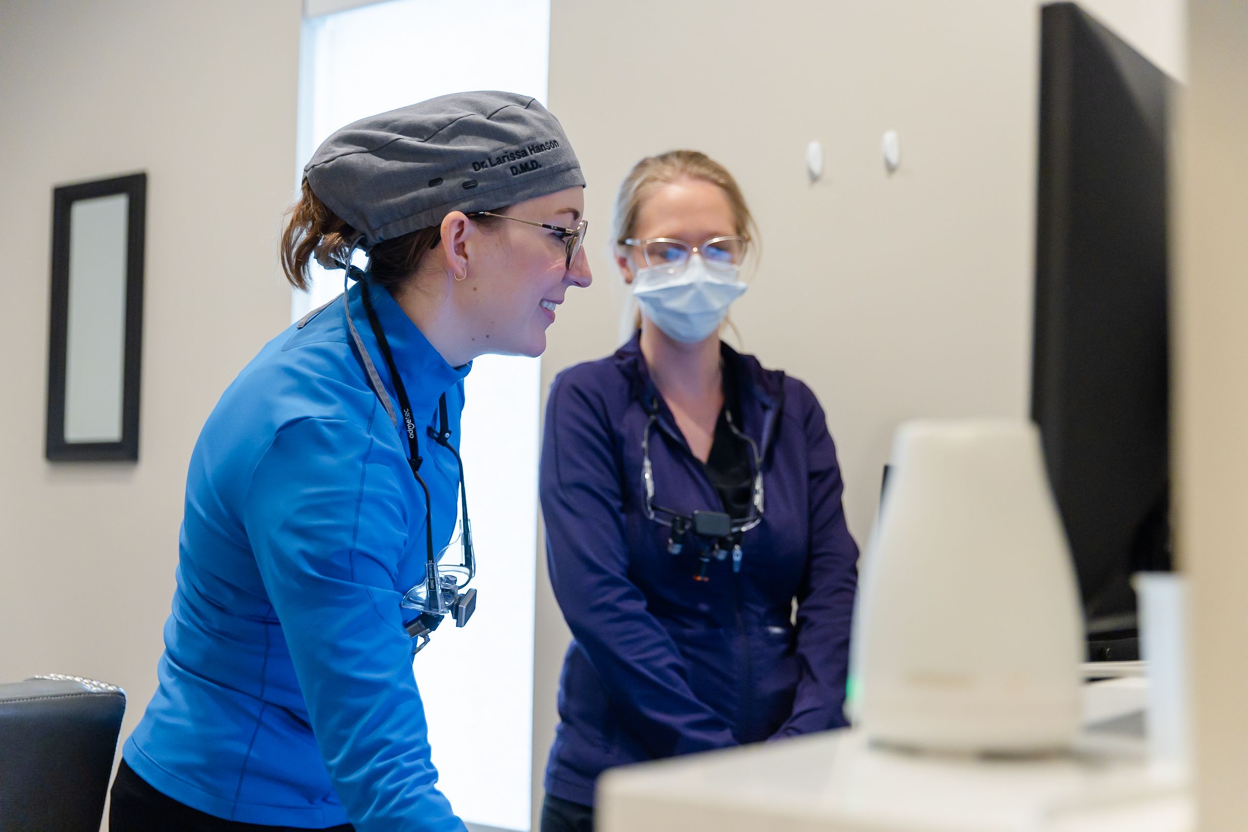 Dr. Larina Mathes in a surgical cap and a colleague with a mask and loupes review a monitor.