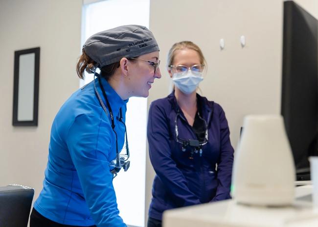 Dr. Larina Mathes in a surgical cap and a colleague with a mask and loupes review a monitor.
