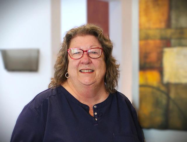 A smiling woman with wavy hair and red-framed glasses wearing a dark blue top.