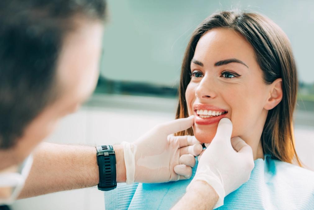 A dentist in white gloves examines a smiling woman's teeth.