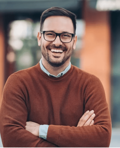 a man wearing glasses and a brown sweater smiles with his arms crossed
