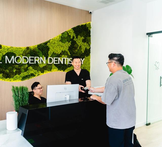 Two smiling staff members interact with a patient at the Bristol Modern Dentistry reception desk, featuring a green moss wall.