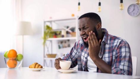 A man grimacing in pain and holding his cheek while holding a coffee cup at a table.