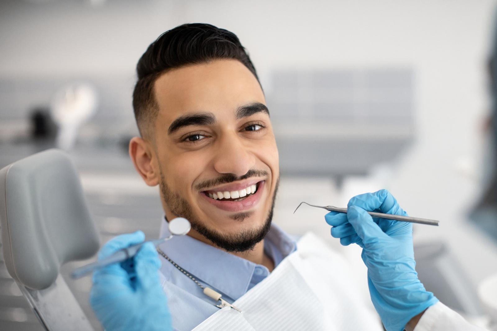 A man with a wide smile in a dental chair, with gloved hands holding dental tools.
