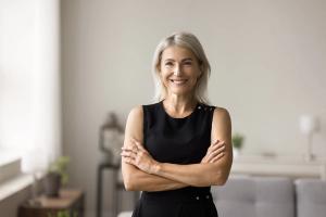 Smiling mature woman with blonde hair in a black dress, arms crossed.