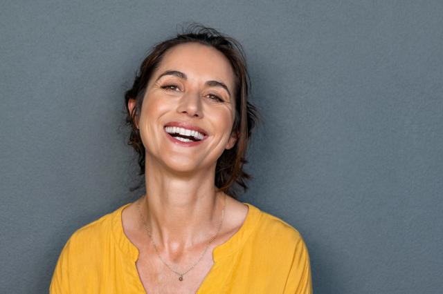 A woman with brown hair laughs brightly, wearing a yellow shirt.