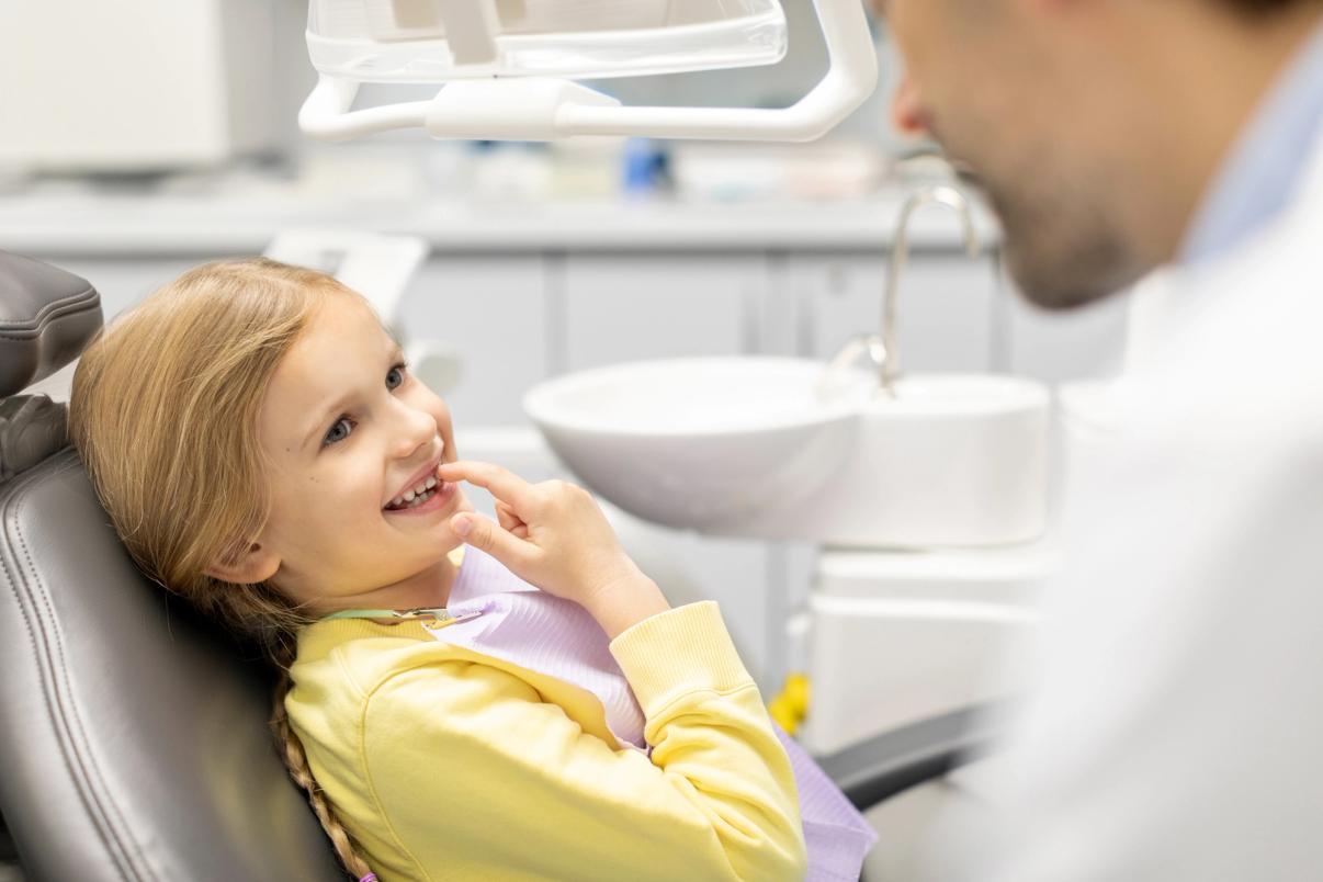 Smiling young girl in a dental chair, pointing at her teeth while looking at the dentist.