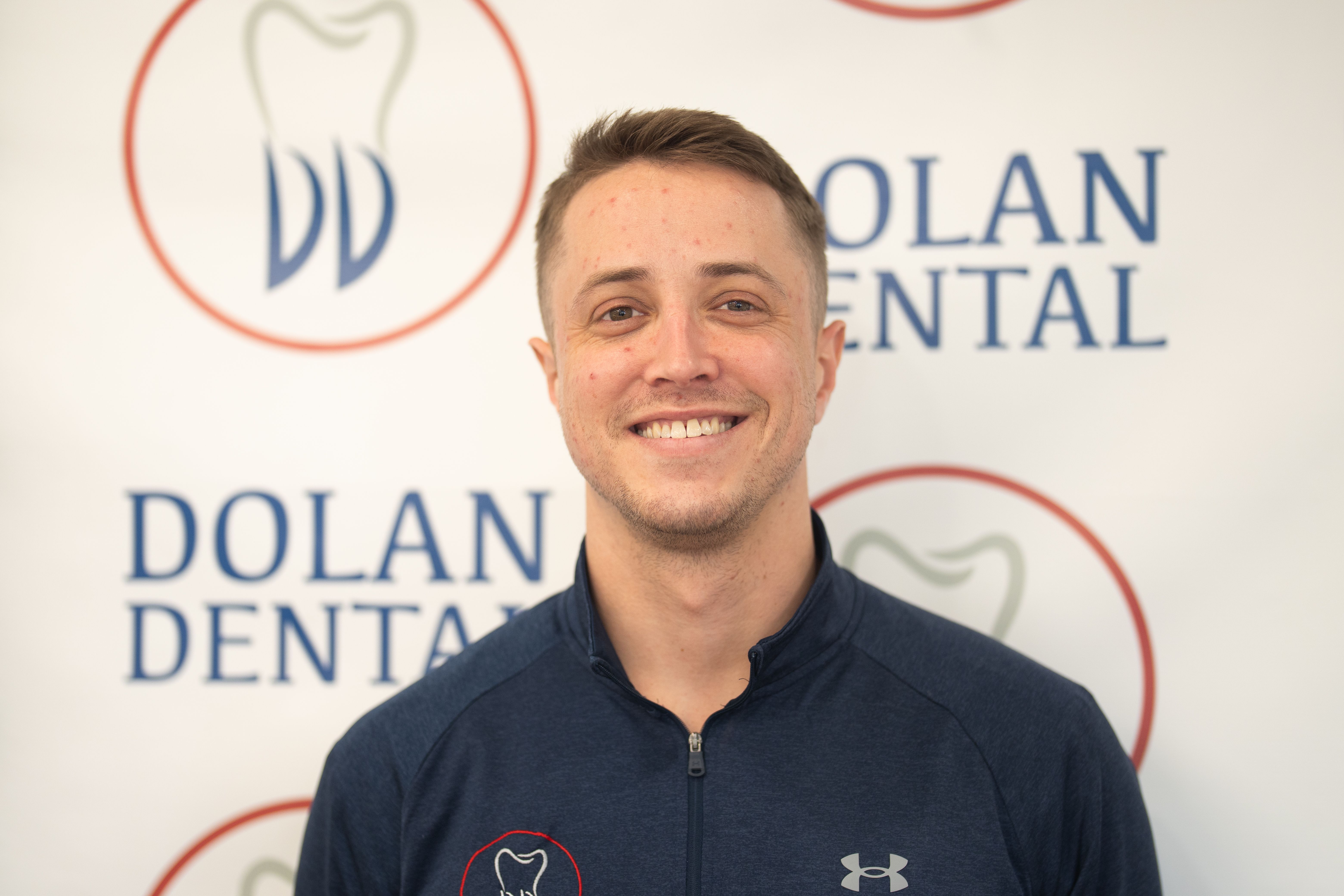 Smiling man wearing a blue athletic top with the Dolan Dental logo, against a branded background.