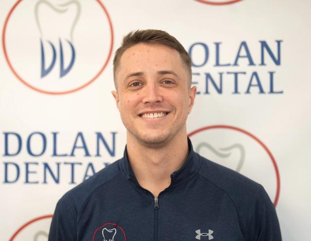 Smiling man wearing a blue athletic top with the Dolan Dental logo, against a branded background.