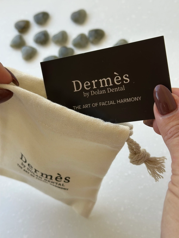 A hand places a black "Dermès by Dolan Dental" card into a beige drawstring bag, with heart-shaped stones blurred in the background.