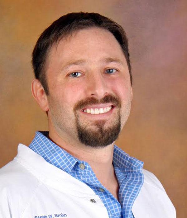 Smiling man with brown hair and a beard, wearing a white lab coat with "Dakota J. Peacher" embroidered on it.