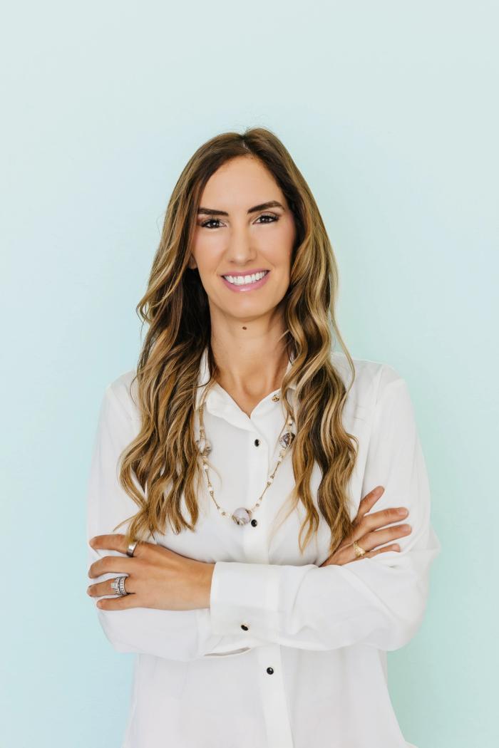 A smiling woman with long wavy brown hair, wearing a white shirt and necklace, stands with her arms crossed against a light blue background.
