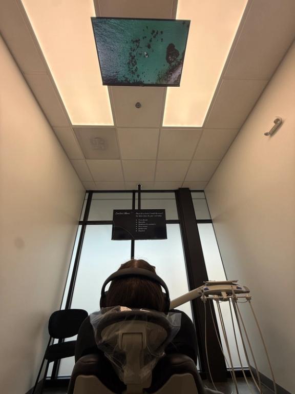 A person in a dental chair with headphones looks up at a ceiling-mounted TV showing an aerial view of turquoise water.