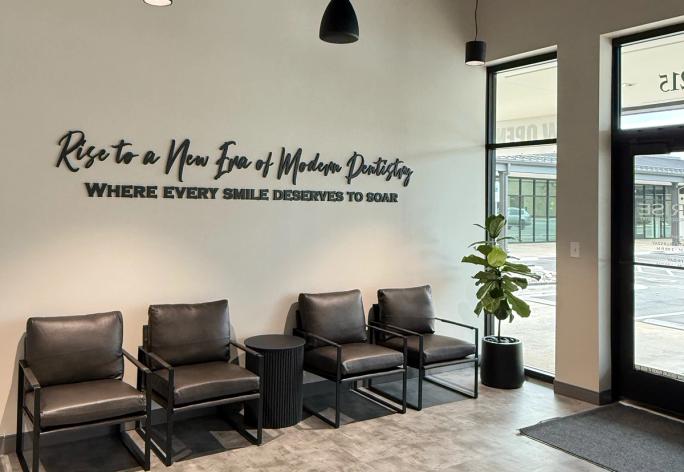 A modern and sleek waiting room with four dark armchairs, a tall green plant, and a wall featuring 3D text that reads "Rise to a New Era of Modern Dentistry WHERE EVERY SMILE DESERVES TO SOAR."