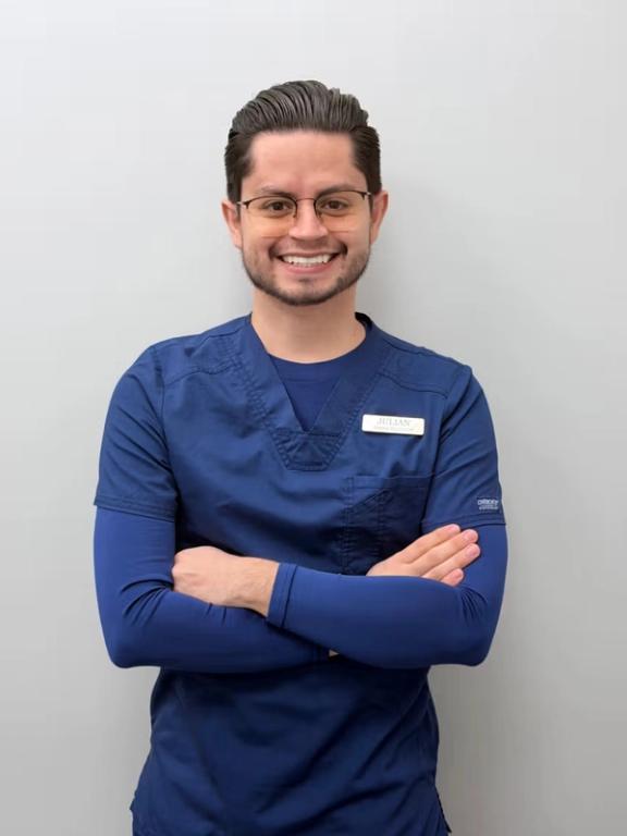 Smiling man Julian in blue scrubs and glasses, arms crossed.