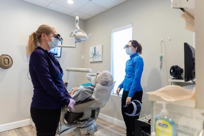 Two masked dental professionals wearing loupes attend to a patient in a dental chair.