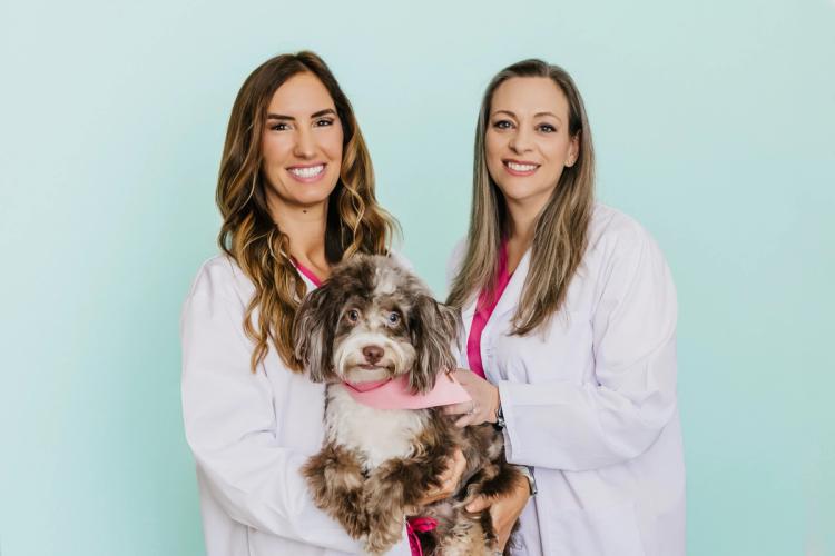 Two smiling women in lab coats and pink scrubs hold a fluffy dog with a pink bandana.