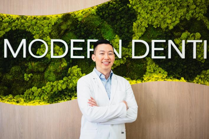 Smiling man in a white lab coat in front of a green moss wall with "MODERN DENTI" in white letters.