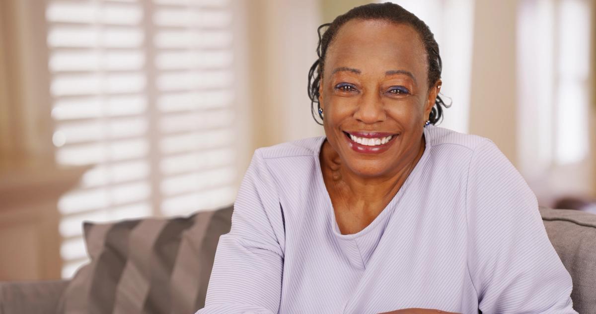 An older Black woman with braided hair smiles broadly while sitting on a couch.