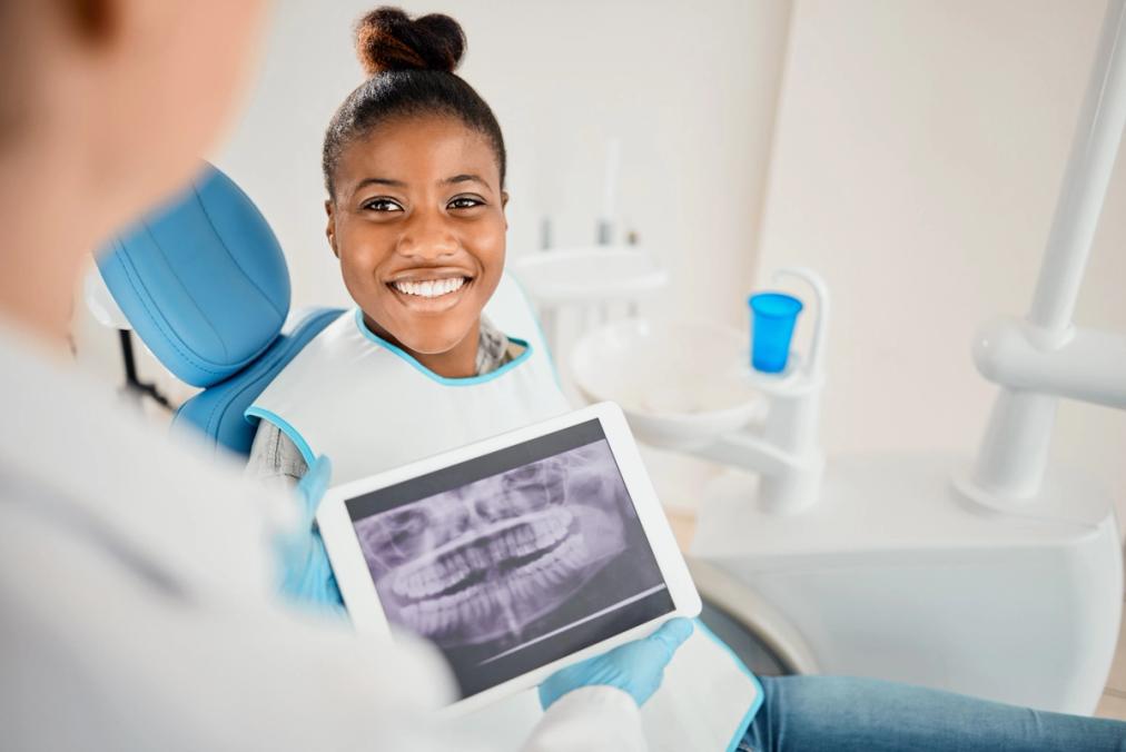 A happy young Black woman in a dental chair looks at a dental professional holding a tablet with her dental X-rays.