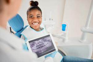 A smiling young Black woman in a dentist's chair looking at a tablet held by a dentist displaying her dental X-rays.