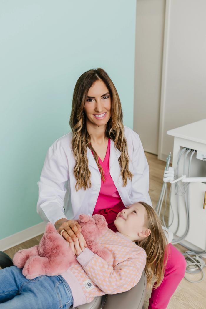 A smiling female dentist comforts a young girl holding a pink stuffed animal in a dental chair.