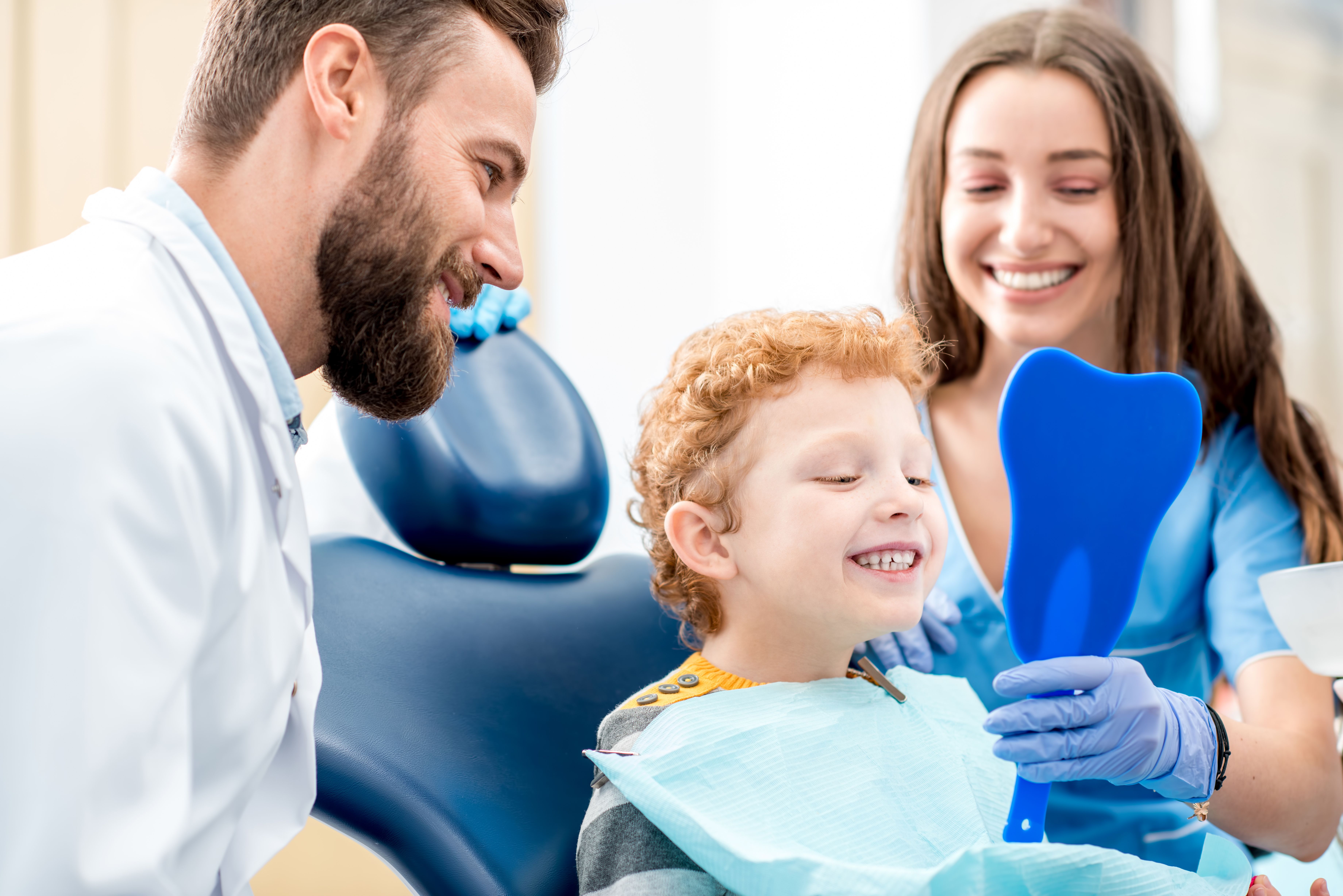 A smiling child looks at their teeth in a blue, tooth-shaped mirror held by a dental assistant, with a male dentist smiling nearby.