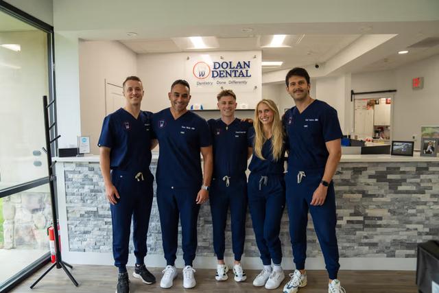 Five smiling dental staff members, four men and one woman, in navy scrubs, stand in front of the Dolan Dental reception desk.