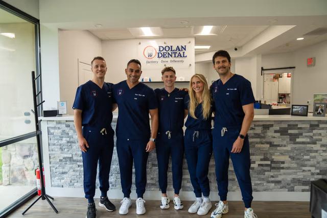 Five smiling dental staff members, four men and one woman, in navy scrubs, stand in front of the Dolan Dental reception desk.