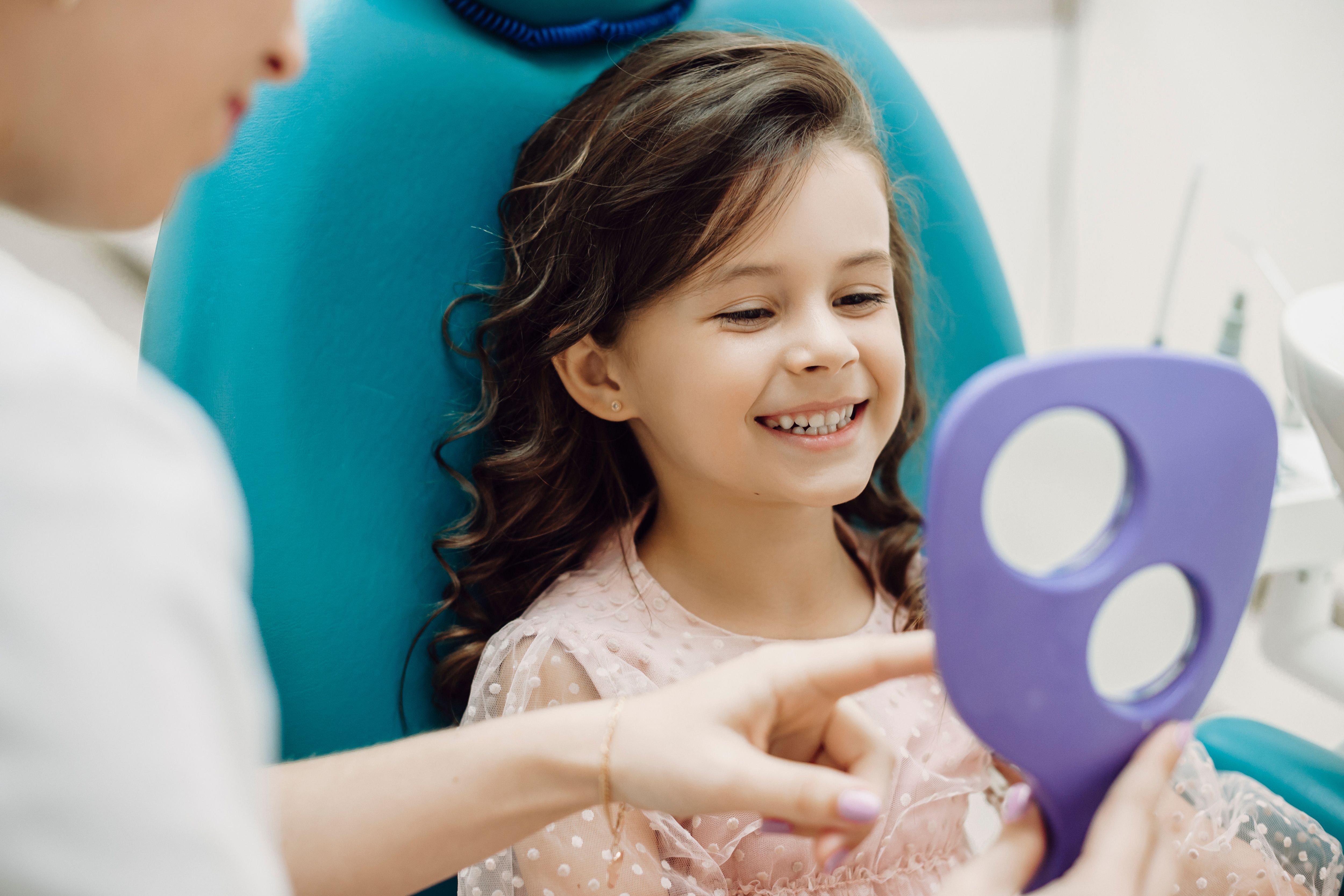 A smiling young girl in a dental chair looking at her reflection in a mirror held by a dental professional.