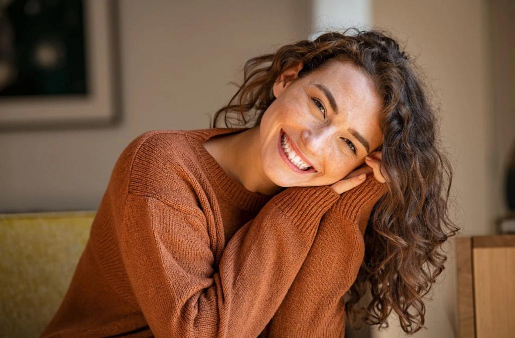 a woman in a brown sweater is smiling while sitting on a couch .