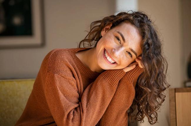 A smiling woman with curly hair and freckles rests her head on her hand.