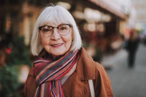 Smiling older woman with white hair and glasses, wearing a brown coat and striped scarf, on a blurred street.