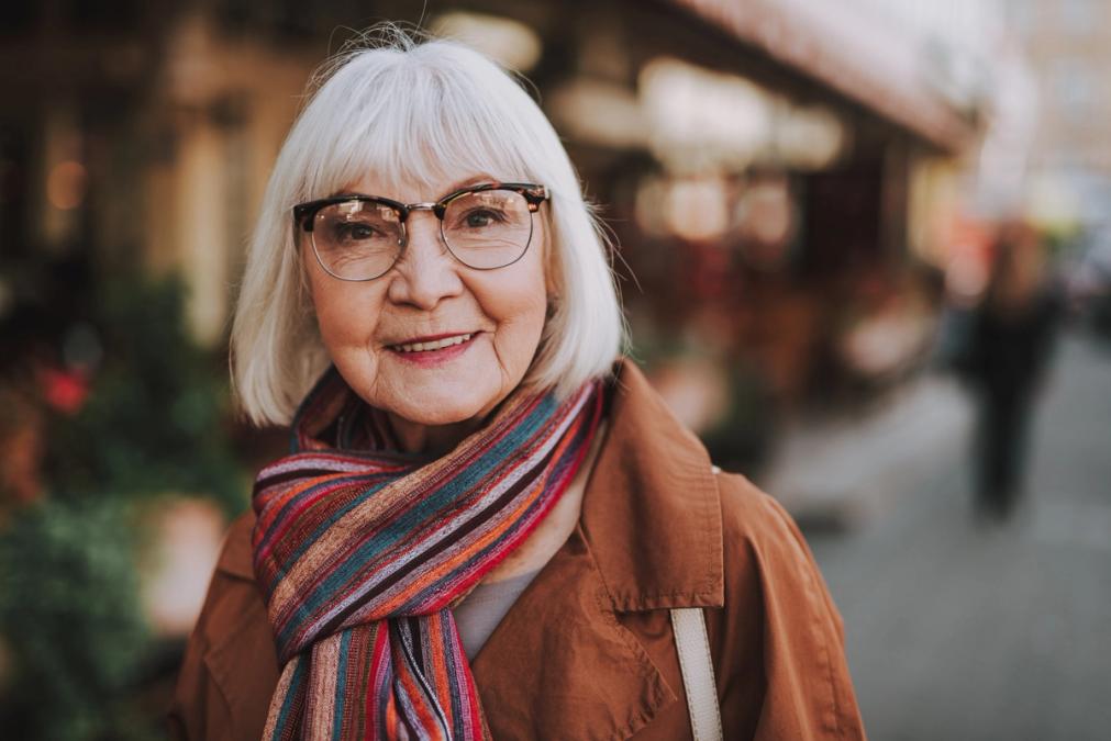 Smiling older woman with white hair and glasses, wearing a brown coat and striped scarf, on a blurred street.